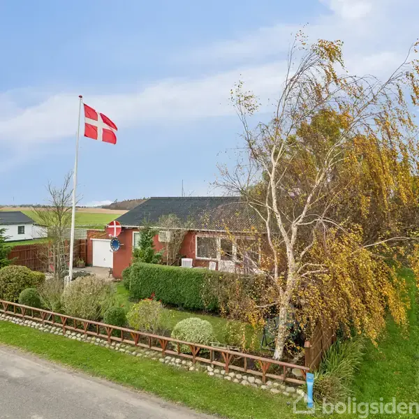 Hus med én etage har en dansk flagstang i haven, omgivet af buske og et træ. Huset ligger ved en vej i et landligt område med marker i baggrunden.