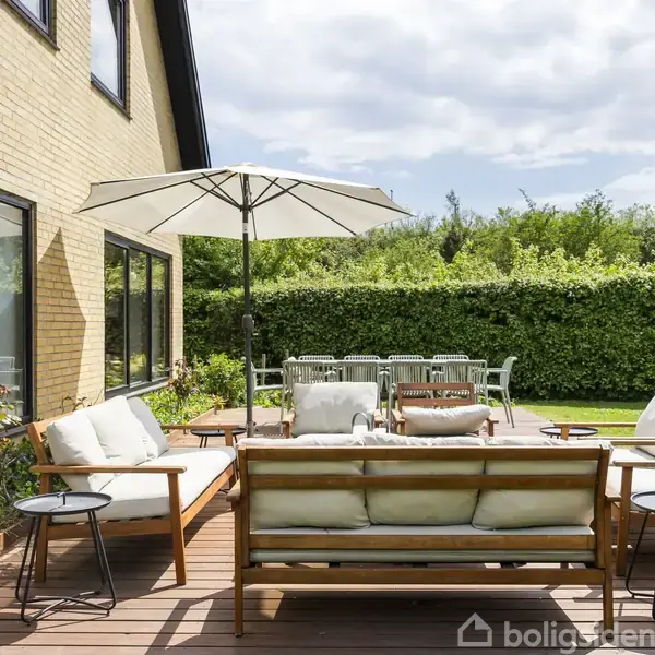 Terrassemøbler med sofagruppe, to stole, borde og en parasol står på en træterrasse ved siden af et hus. I baggrunden ses en stor have omgivet af grønne buske.