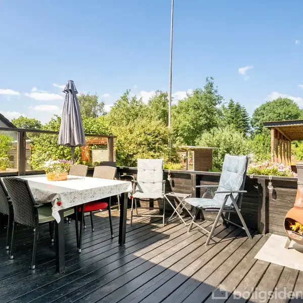 En terrasse med sorte trægulve. Havebord med stol omkring, en parasol og grill i hjørnet. Omgivet af grøn beplantning under en klar blå himmel.