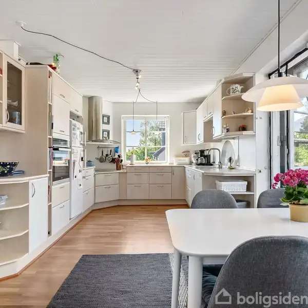 Et køkken med åbne hylder og skuffer og et indbygget ovn-tårn. Der står et spisebord med grå stole og en potteplante. Vinduerne giver et syn til en have.