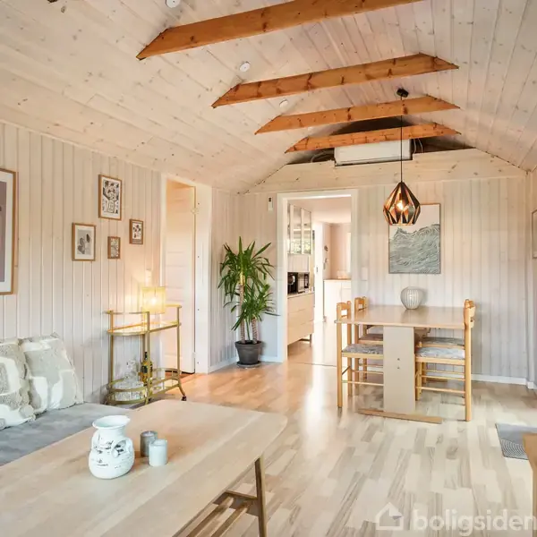 Stue i lyst træ med synlige loftsbjælker. Langbord med fire stole står centralt. Sofa med puder til venstre. Lampe hænger over bordet. Døre fører til køkken.