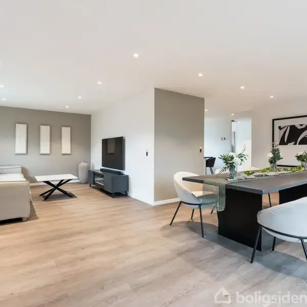 Stue med lysegrå sofaer, fladskærms-tv på mediemøbel, og spisebord med seks hvide stole til højre. Moderne kunst på væggene, stort vindue og trægulv.