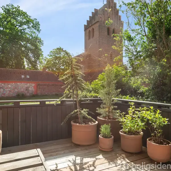 En terrasse med flere planter i potter står stille. I baggrunden ses en kirke med klokketårn omgivet af træer. Solen skinner på den rolige, grønne have.