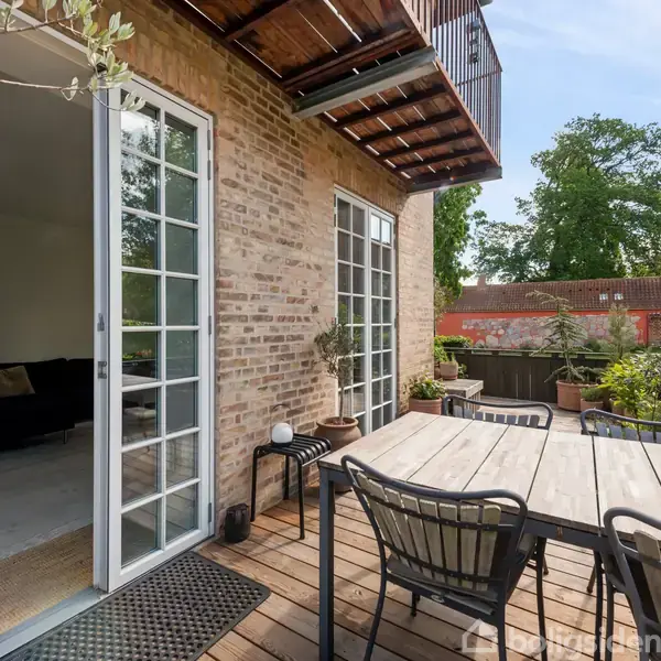 Terrasse med træbord og stole i sollys. Dør i glas fører til stue i bygning af mursten. Omkring er der grønne planter og rødt tag i baggrunden.