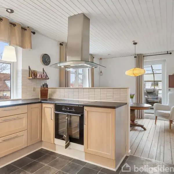 Moderne køkken med en kogeø, indbygget ovn og emhætte; det støder op til en spisestue med runde bord og komfortable stole. Store vinduer lader naturligt lys ind. Ingen tekst.