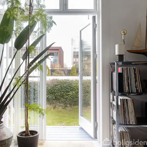 En åbnet glasdør fører fra en stue ud til en grøn have. En reol med bøger står til højre, og en stor plante ses til venstre.