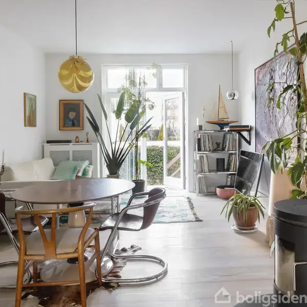 Et moderne dagligstueinteriør med et rundt spisebord og stole. Rummet er lyst med grønne planter, sofagruppe, reoler og stor kunst på væggen. Sollys strømmer ind gennem store vinduer.