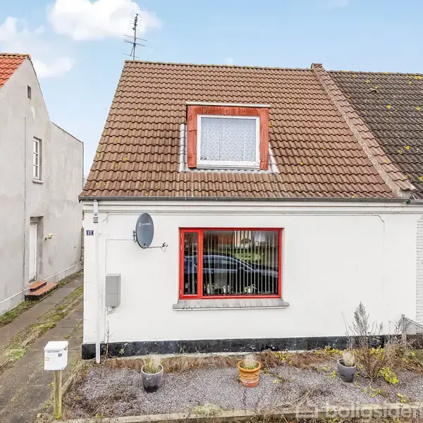 Hus med rødtag udgør hovedobjektet; forgrunden viser en have med potteplanter, og huset har en hvid facade med rødt, gitterindrammet vindue. Beliggende på en smal gade med tilstødende bygning.