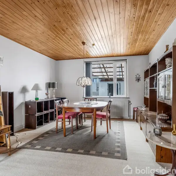 Spisebord med stole står i midten af en stue. En pendellampe hænger over bordet. Stuen har et træloft, reoler langs væggen og et vindue mod en terrasse.