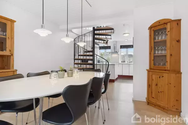 Et ovalt, hvidt spisebord med sorte stole står i et lyst rum med en trætrappe. I baggrunden ses et køkken. Skabe af træ er placeret i hjørnerne.