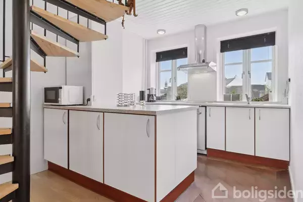 Et moderne køkken med hvide skabe og mørke bordplader. En mikrobølgeovn står på disken. En spiraltrappe er synlig til venstre, og der er to vinduer i baggrunden.