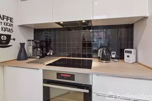 En moderne køkkenbordplade med en indbygget kogeplade; en kaffemaskine, elkedel og brødrister står ovenpå. Sort flisebelagt væg i baggrunden. Tekst: "KAFFE KRAM KRUS".