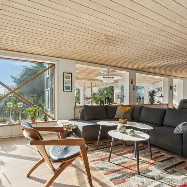 En moderne stue med en stor hjørnesofa i sort, placeret på et farverigt tæppe. Store vinduer giver masser af lys, og planter står ved vindueskarmen. Træloft og gulv tilføjer varme.