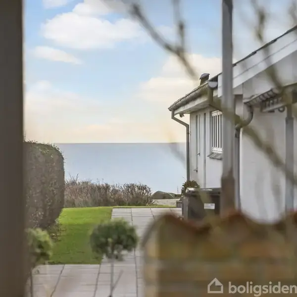 Hus i forgrunden, står stille, omgivet af en flisebelagt terrasse og grønne hække, med udsigt til horisonten over en rolig havoverflade under en blå himmel med få skyer.