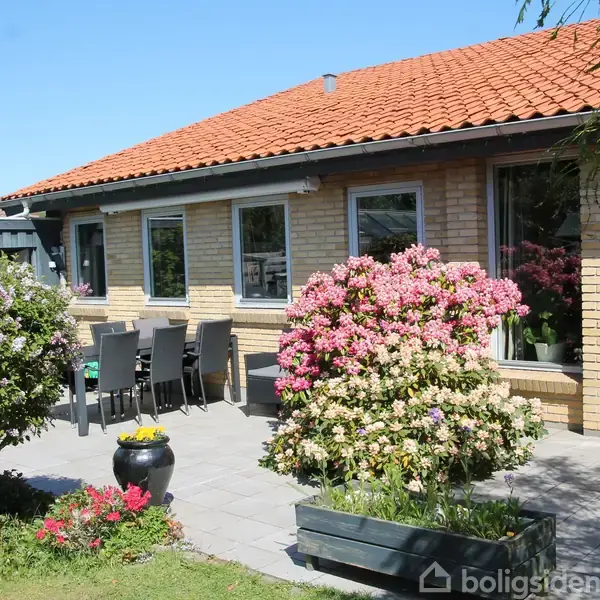Et gulstenshus med rødt tegltag har en terrasse med havemøbler. Pink og hvide buske blomstrer i plantekasser og tæt ved, med en klar blå himmel i baggrunden.