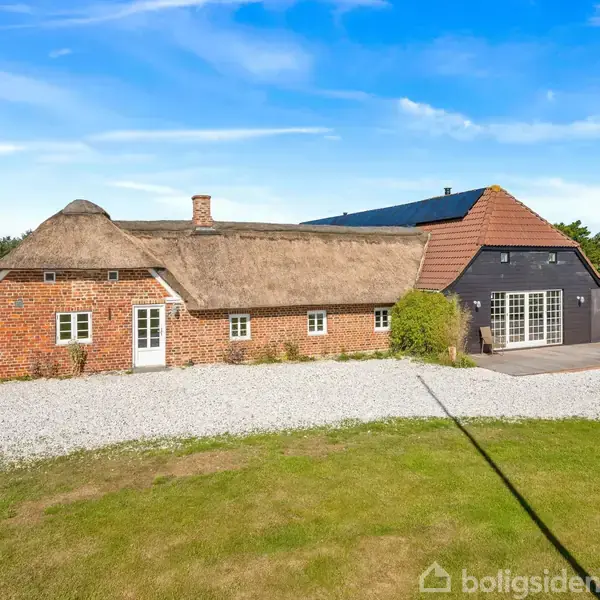 En traditionel, stråtækt murstensbygning med flere små vinduer står stille på en grusbelagt gårdsplads, omgivet af en grøn græsplæne og træer under en klar blå himmel.