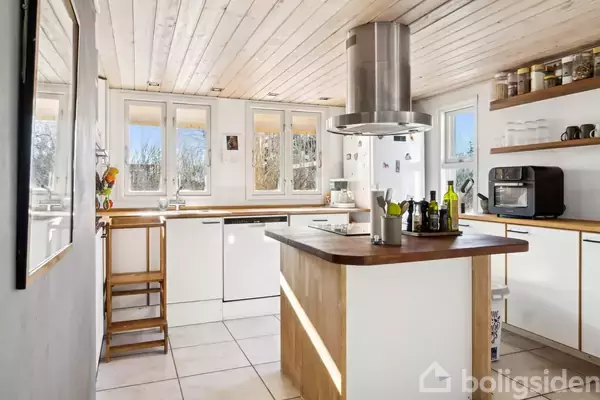 Et moderne køkken med en central køkkenø indeholder flasker og krydderier, omgivet af hvide skabe og store vinduer, der slipper naturligt lys ind. Hylder med kopper og mikroovn ses.