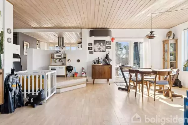 Stue med trægulve, træloft og store vinduer. Legetøj er placeret ved en lille låge. Spisebord og stole står ved siden af et vitrineskab. Hyggelig og lys atmosfære.