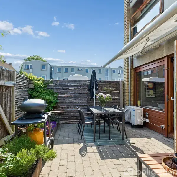 En terrasse med flisebelægning har et havebord og stole under en markise. En grill står til venstre blandt planter. Bagved ses en træhegn og flere bygninger i baggrunden.