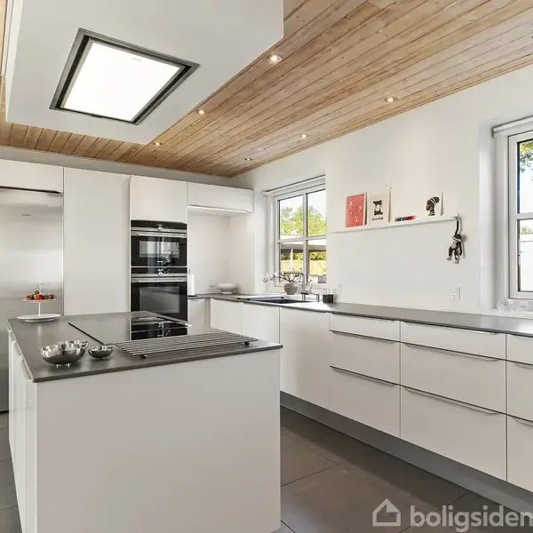 Et moderne køkken med hvide skabe og en central ø med kogeplade. I baggrunden ses to store sølvfarvede køleskabe. Loftet er træ, og der er vinduer mod en have.