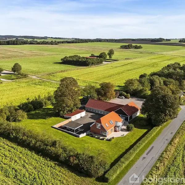 En landejendom med rødt tag omgivet af grønne marker og træer. Ejendommen ligger ved en landevej i et åbent landdistrikt.