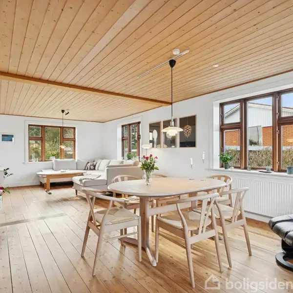 Et spisebord med stole står i midten af et lyst, træbeklædt rum. Til venstre ses en sofa ved vinduer. På bordet står en vase med blomster.