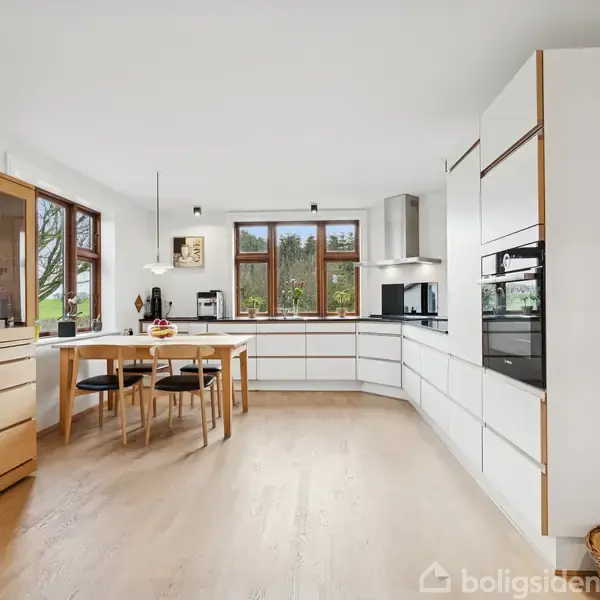 Et lyst køkken har et træpanel-sæt med et spisebord og fire stole i centrum. Skabs- og køkkenapparater er i hvide nuancer. Vinduer giver udsigt til en grøn have.