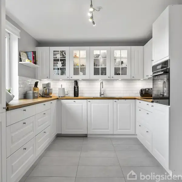 Et hvidt køkken med træbordplader omfatter skabe, ovn og vask; lysefliser langs væggen, loftsbelysning. En kalender hænger til venstre. Moderne og ryddeligt interiør.