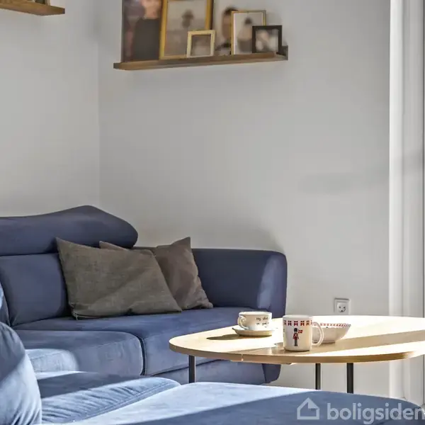 Blå sofa med puder står ved siden af et rundt træbord med kaffekopper. Bagved hænger hylder med billeder på en hvid væg, ved siden af en stor glasdør til en terrasse.