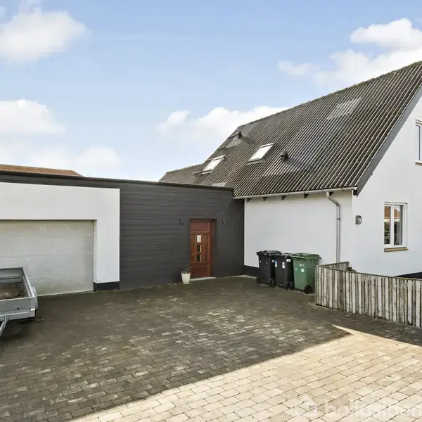 Et moderne, hvidt hus med skråt tag står stille ved en indkørsel med fliser og trailer. Til højre er der en lille have og skraldespande ved væggen.