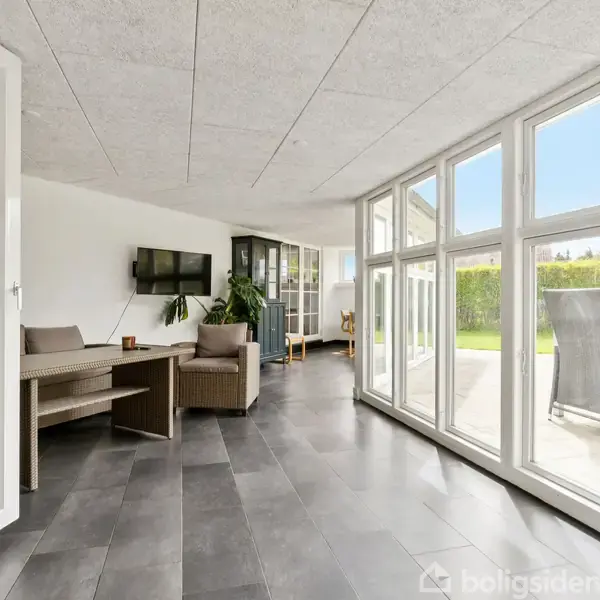 Franske døre åbner til en lys stue med grå fliser, sofaer, og et TV på væggen. Store vinduer giver udsigt til en terrasse med havemøbler i grønne omgivelser.