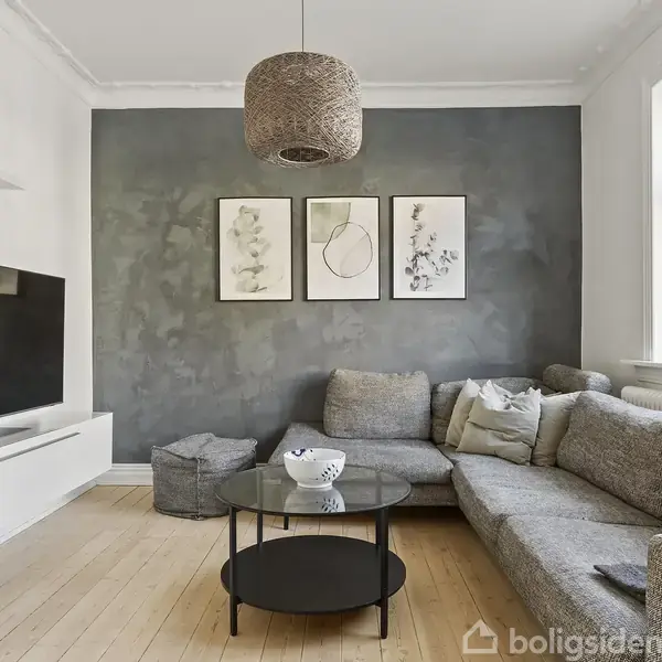 En grå sofa står i en moderne stue med trælaminatgulv. Væggene har kunsttryk og en dæmpet farvetone. Et rundt, sort bord er placeret foran sofaen, og et fladskærms-TV er monteret på den modsatte væg.