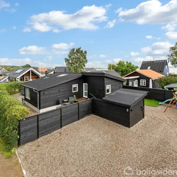 Sort hus med fladt tag, omgivet af grusbelagt gårdsplads, indhegnet af sort stakit. Gyngestativ i baggård. Nabohuse og grønne områder ses i baggrunden under en blå, skyet himmel.