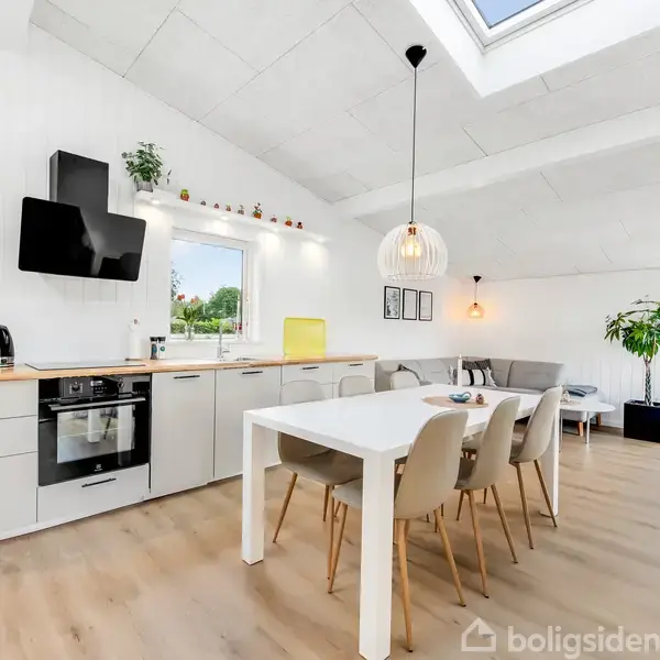 Et lyst, moderne køkken-alrum med hvidt loft og trægulv. Et spisebord med fire stole står i rummet. Køkkenet har hvide skabe, komfur og emhætte. En stue i baggrunden.