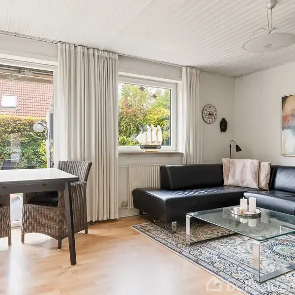 Sort sofa ved glasbord med lys, placeret på tæppe. Rattan spisebord med fire stole til venstre. Store vinduer med hvide gardiner, have udenfor. Moderne vægkunst til højre.