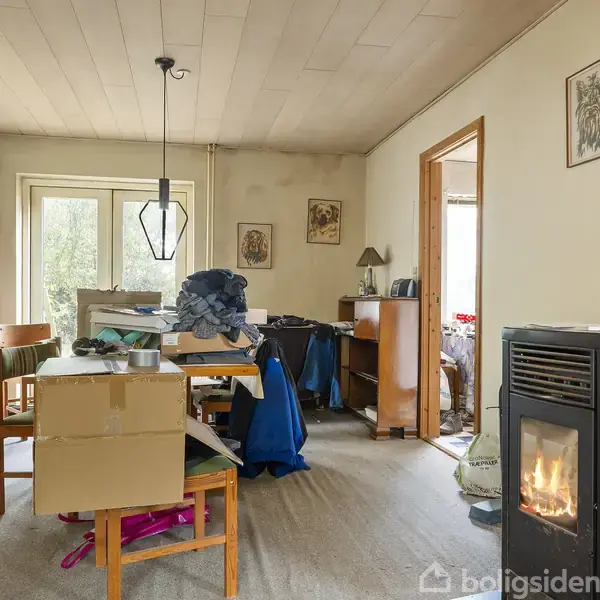En brændeovn opvarmer et hjemmerum fyldt med møbler og kasser. Der er en lampe hængende fra loftet, og vinduer lader naturligt lys strømme ind i rummet.
