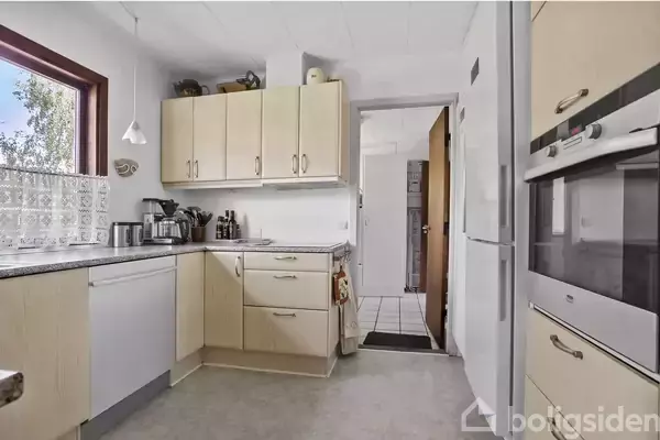 Køkken med lyse skabe og skuffer, moderne apparater, opvaskemaskine og kaffemaskine på bordpladen. Vindue til venstre med udsigt til træer. Dør fører til anden rum.