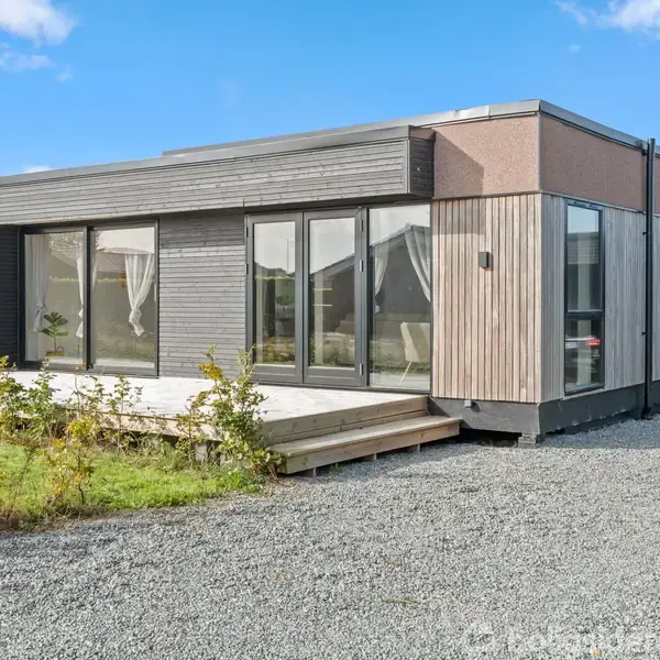 Et moderne, enkelt hus står på en grusvej omgivet af græs og buske. Store vinduer pryder facaden, hvilket giver et lyst udtryk mod en klar blå himmel.