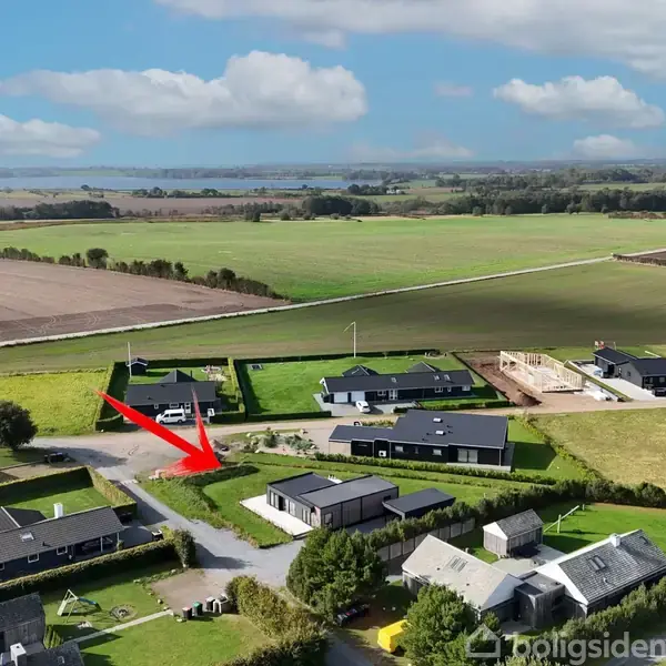 En rød pil peger mod et hus i en landlig landsby. Omgivet af andre huse, marker og en fjern sø under en blå himmel med skyer.