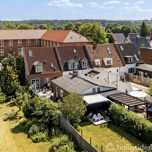 Rækkehuse med røde tage står langs en velplejet have, hvor der er en pergola og havemøbler. Bagved ses en stor bygning og frodige træer under en klar blå himmel.