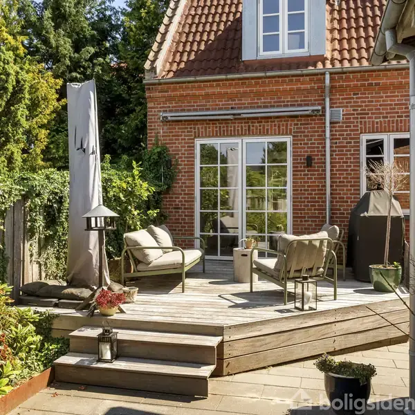 Terrasse med træmøbler, parasol og lanterne nær rødt murstenshus. Trædæk med planter omgivet af træhegn og grønne buske. Husets tag er rødt med hvide vinduer.