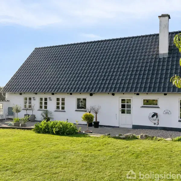 Et hvidt hus med sort tag står på en velplejet græsplæne omgivet af buske og træer. Huset har store vinduer og en terrasse på siden.