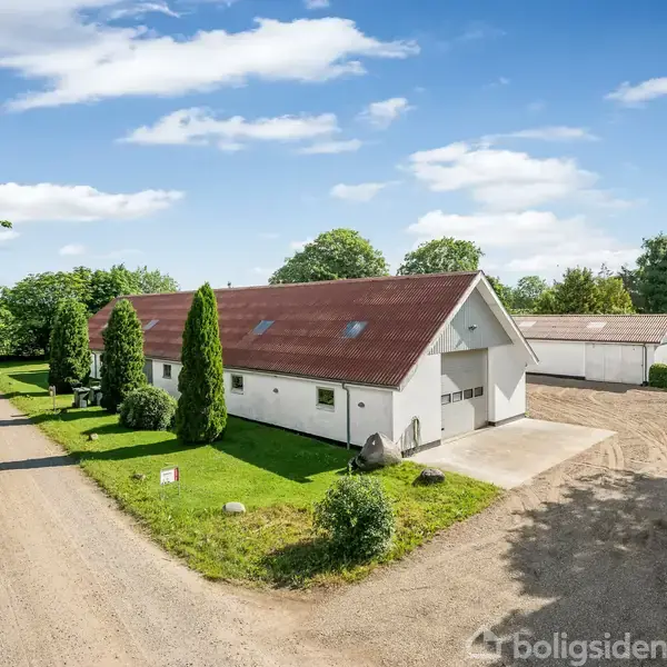 En hvid bygning med rødt tag står ved en grusvej. Bygningen er omgivet af grønne træer og græs. Det er solskinsvejr med få skyer på himlen.