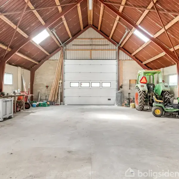 En stor garage med højt trætag og betonbund, indeholdende en traktor og værktøj. Lysindfald kommer fra ovenlysvinduer. Der er opbevaring på venstre væg.