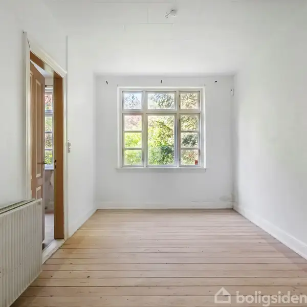 Et tomt rum med trægulv har et vindue, der lukker naturligt lys ind. Til venstre er en radiator og en åben dør. Væggene er hvide.
