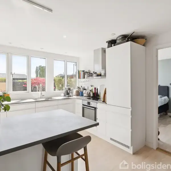 Køkken med hvidt interiør og apparater, set fra en morgenmadsbar med en potteplante. Store vinduer giver udsigt til en have. Døråbning fører til soveværelse.