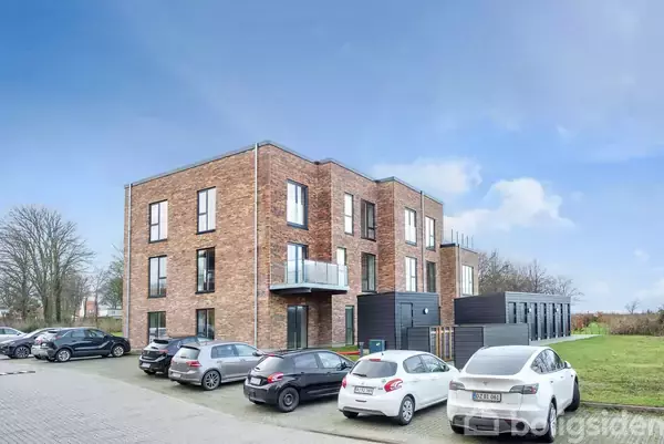 Et tre-etagers murstensbyggeri står stille i en parkeringsplads, omgivet af flere parkerede biler og et grønt område i baggrunden under en blå himmel.