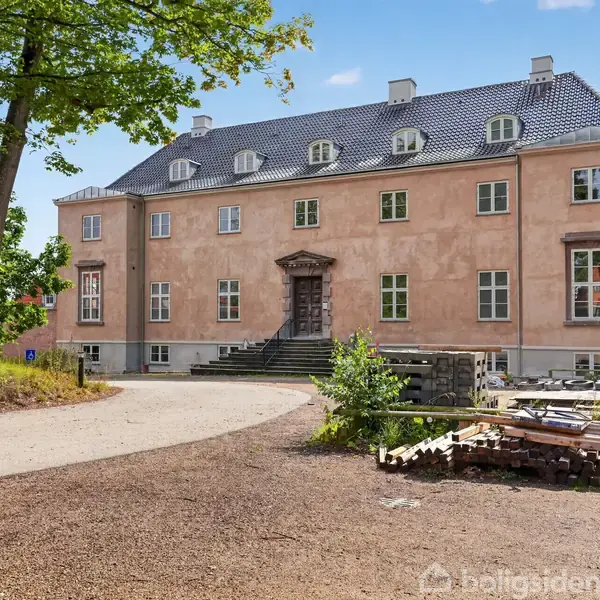 En stor, lyserød bygning med skifertag står omgivet af træer i en åbning. Foran bygningen er der en grusvej og stabler af byggematerialer.