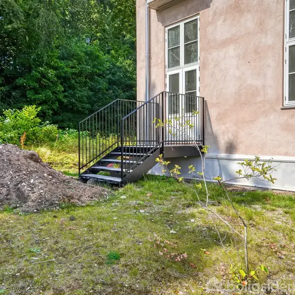 En metaltrappe fører op til en dør på et hus med lyserød facade. Ved siden af trappen er en jordbunke, omgivet af græs og træer i en have.