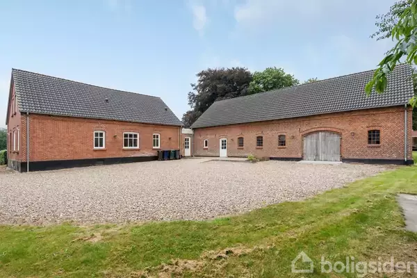 To røde murstensbygninger med skifertage omkranser en grusbelagt gårdsplads. Omkring gården er der grønne græsarealer og træer, som danner en fredelig landskabsramme.